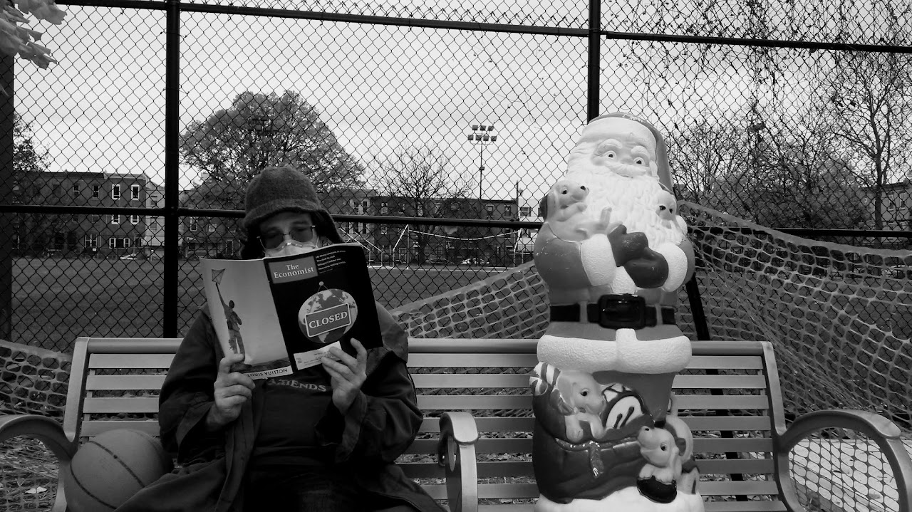 Black-and-white still of a man reading beside a plastic Santa Claus on an empty playground bench